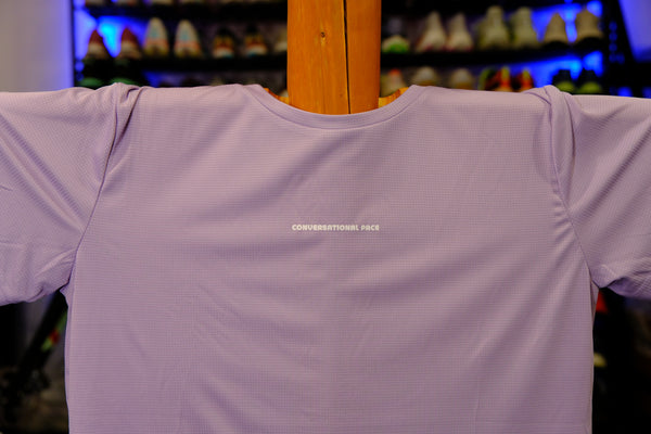 Light purple t-shirt with 'Conversational Pace' text on a wooden hanger against a blurred background.
